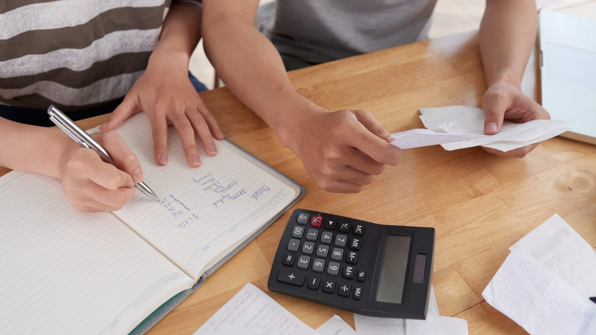 Two people at a wooden table sorting receipts, with one writing figures in a notebook beside a calculator