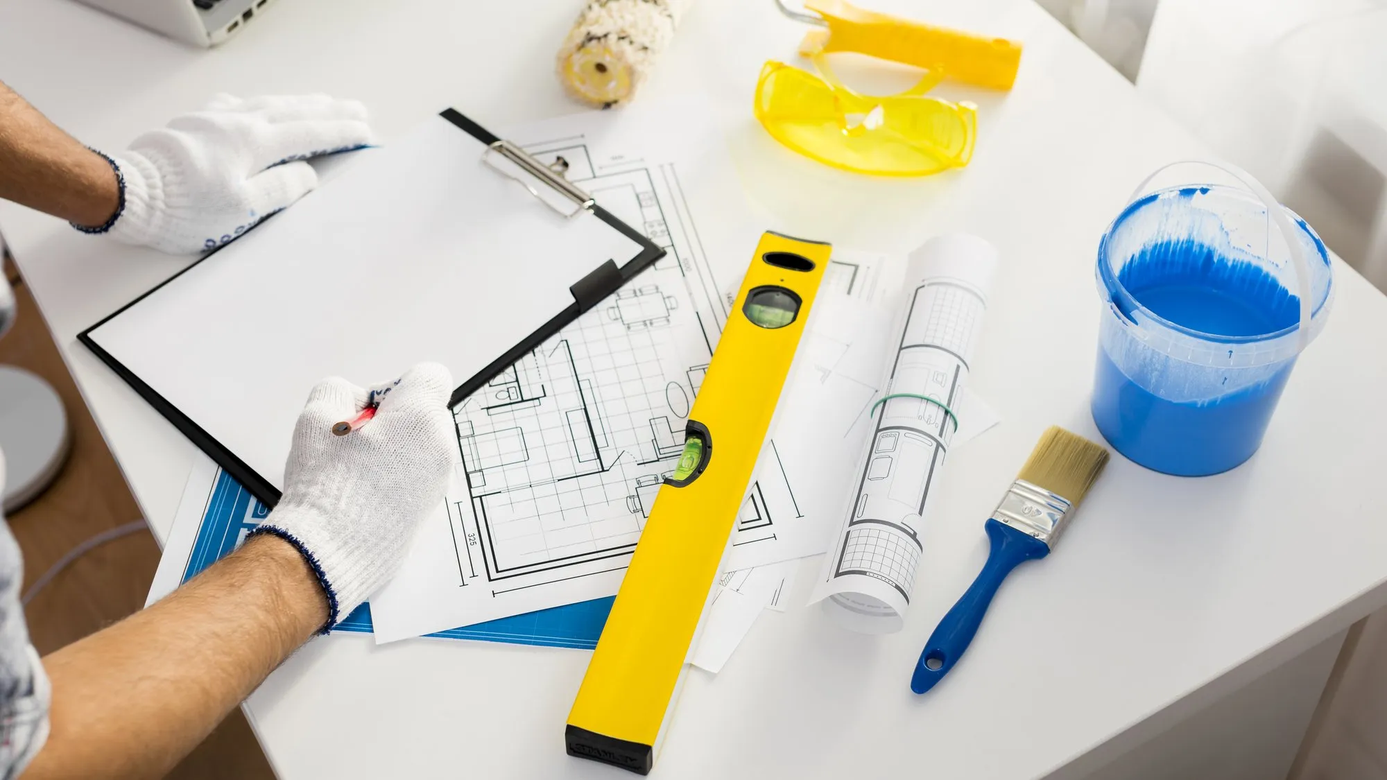 Gloved hands holding a clipboard over house floor plans, next to a yellow spirit level, a paintbrush, a paint roller and a tub of blue paint