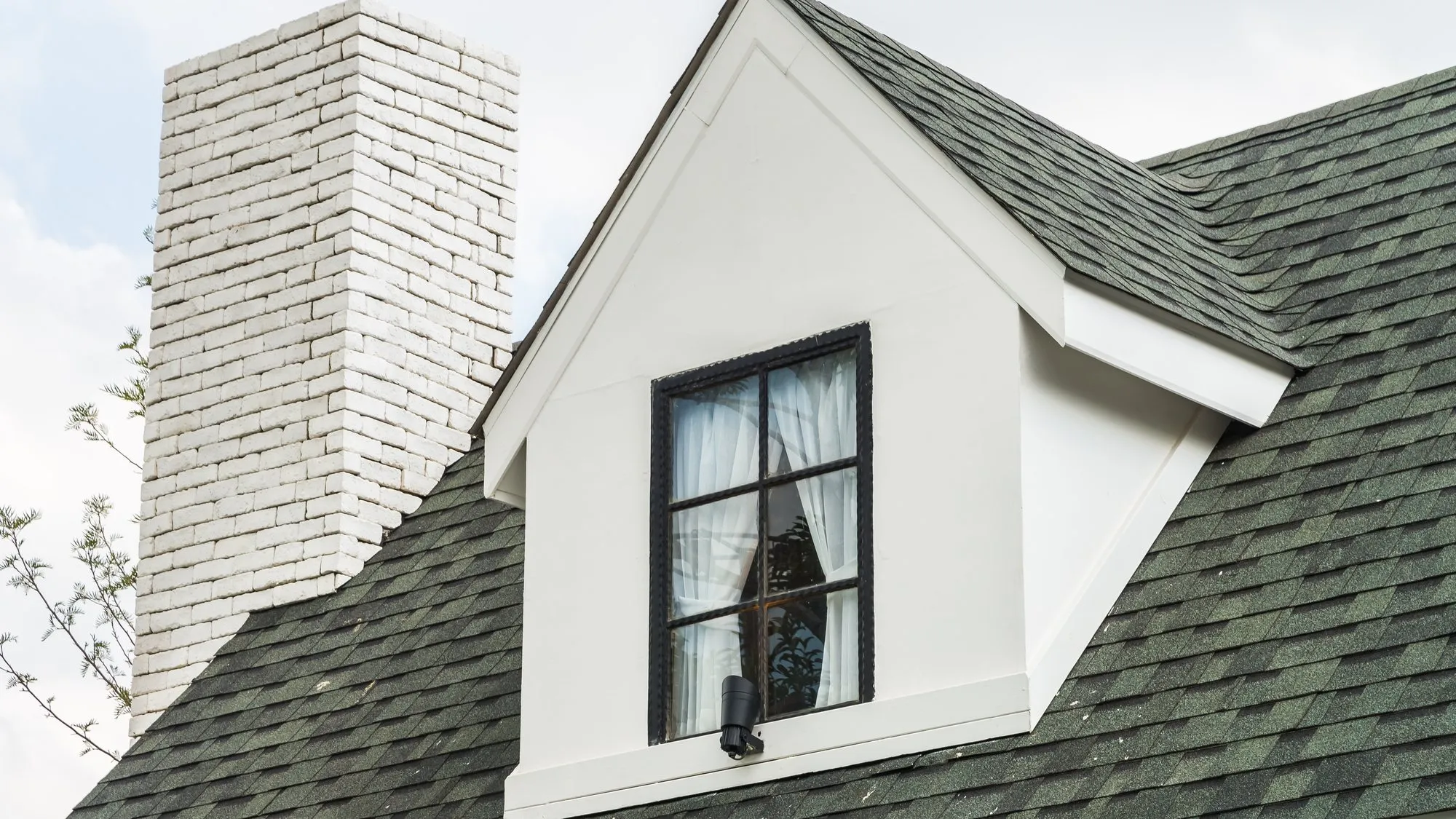 The dark green shingled roof of a house with a white dormer window and a white brick chimney against a cloudy sky