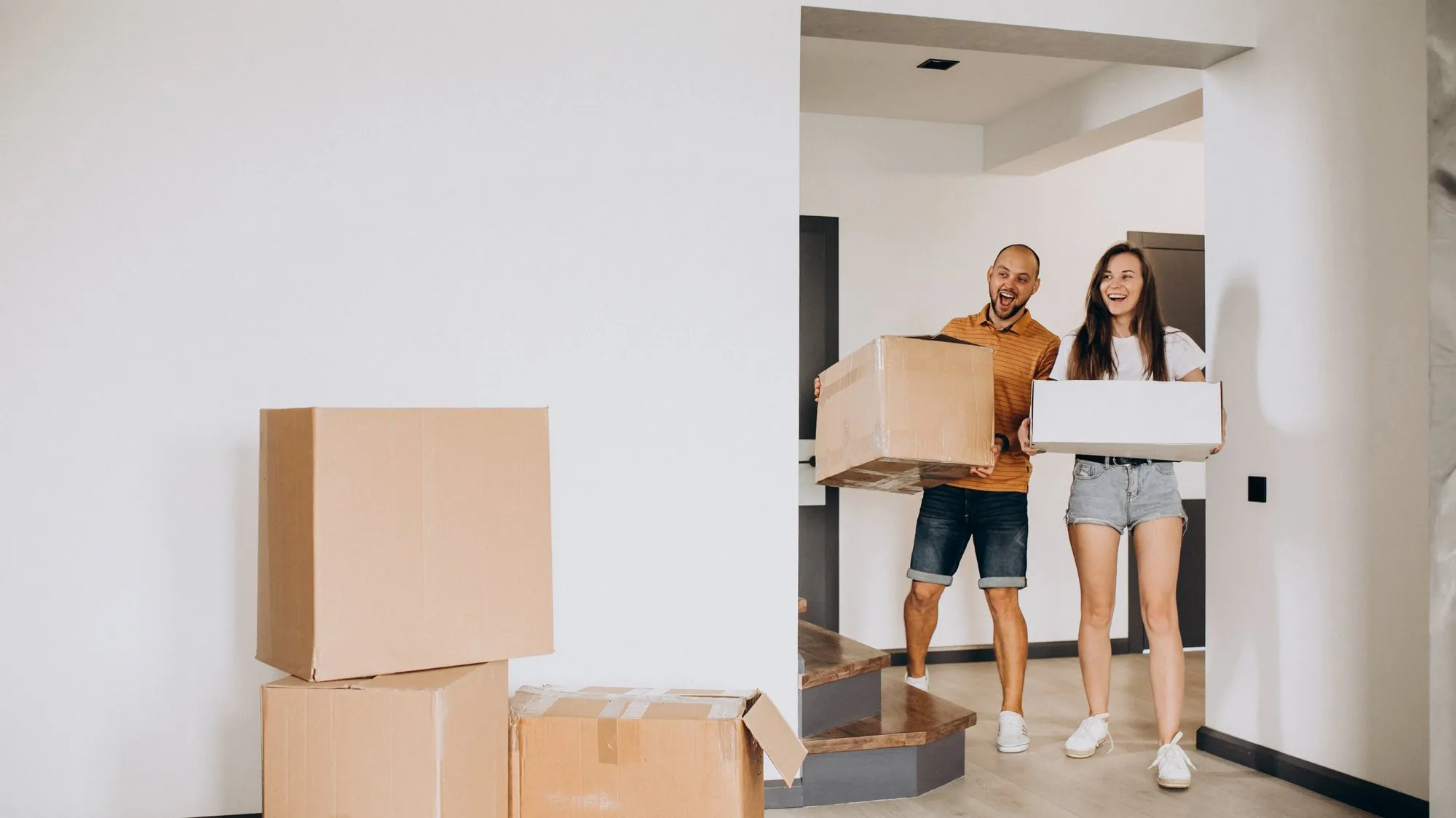 A smiling couple carrying cardboard boxes into an empty, white-walled home, with more boxes stacked in the foreground
