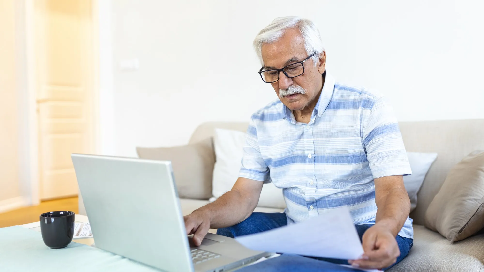 A gray-haired man in glasses sits on a couch reading a sheet of paper while typing on a laptop, with a black mug on the coffee table