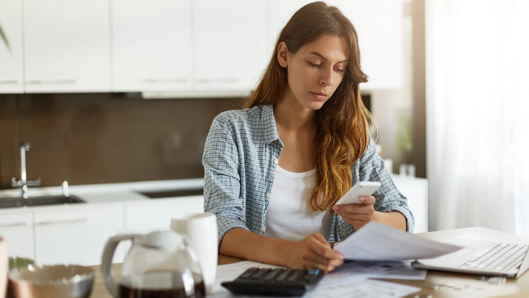 A woman at a kitchen table reading papers and her phone, with a calculator, a laptop and a coffee pot beside her