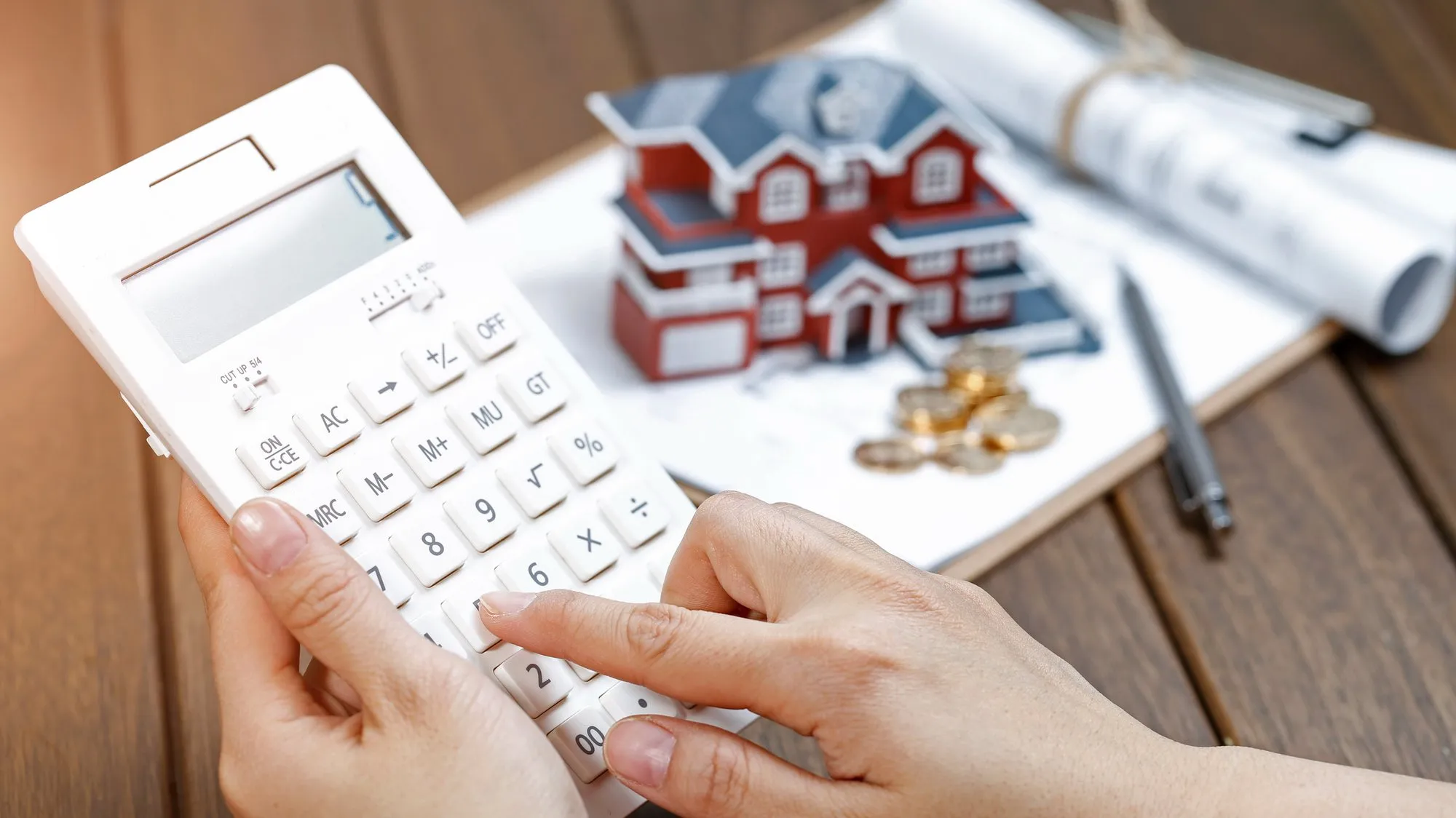 Hands using a white calculator in front of a model house, with coins, a pen and rolled-up plans on a wooden table